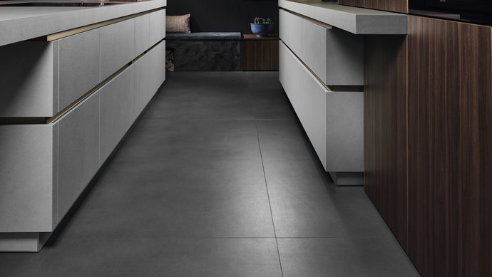 Blick durch eine Inselküche in grauer Strukturoberfläche mit bronzefarbenen Griffmulden und holzfarbenen Hochschränken