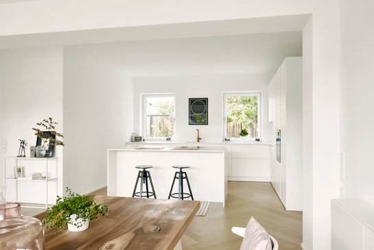 Weiße Inselküche mit Barhockern hinter einer Esstischplatte aus Holz