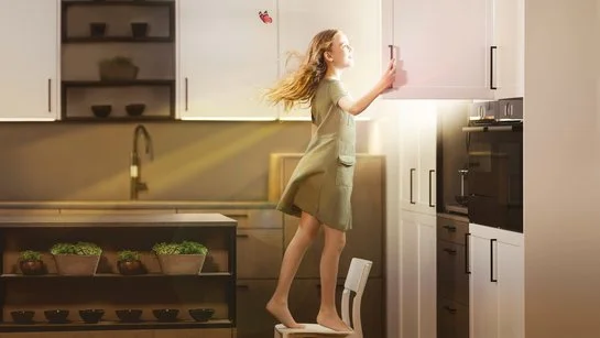 Ein Mädchen steht in einer Küche auf einem Stuhl und öffnet einen Oberschrank, daraus scheint die Sonne.