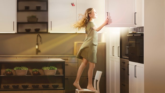 Ein Mädchen steht in einer Küche auf einem Stuhl und öffnet einen Oberschrank, daraus scheint die Sonne.