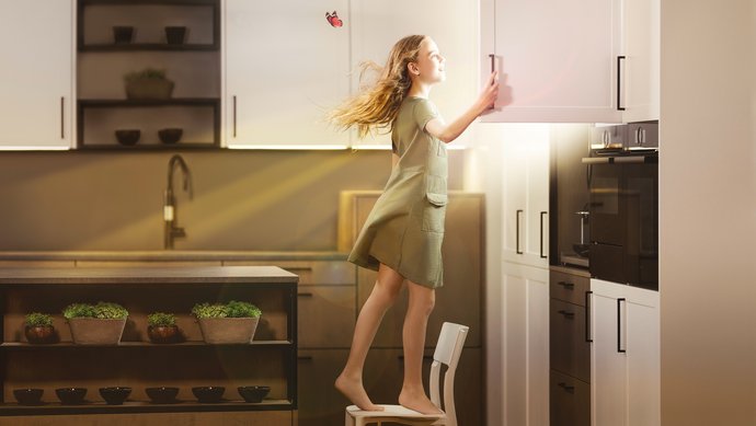 Ein Mädchen steht in einer Küche auf einem Stuhl und öffnet einen Oberschrank, daraus scheint die Sonne.