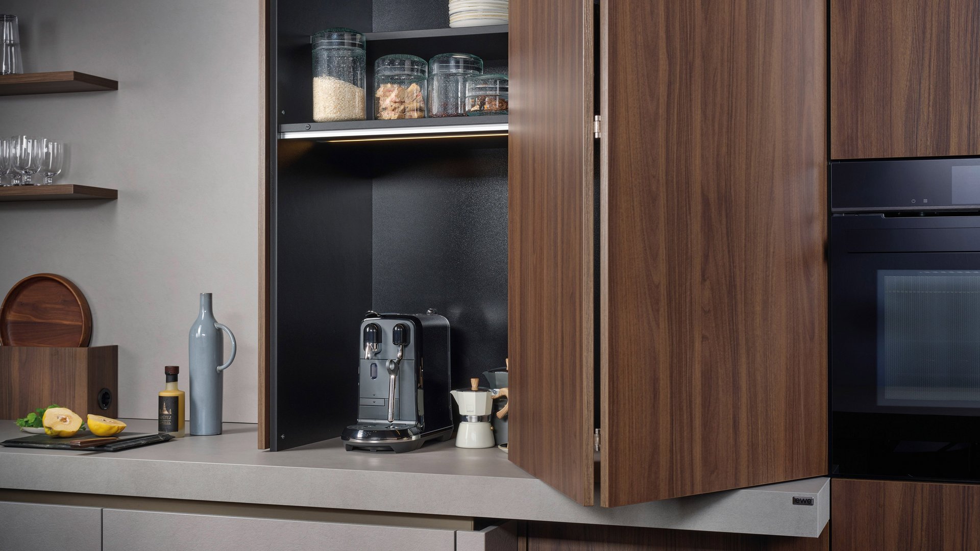 ewe Cemento . ewe Sintesa in malta grigio . nuss pavia, Falttürschrank Halb geöffneter Falttürschrank in Nuss-Optik in einer sonst grauen Küche